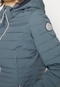 Teal puffervinterjakke med lodret quiltning, en hætte med snoretræk og et rundt logo-patch på venstre ærme. Har en lynlåslukning foran.