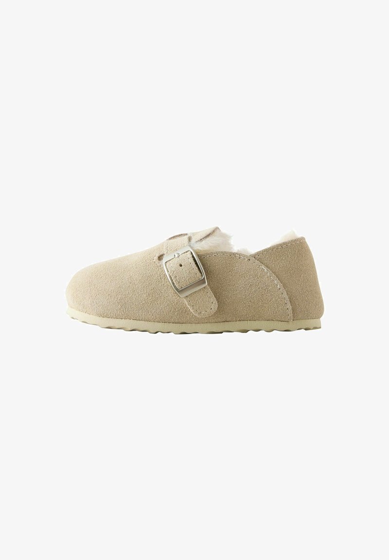 Chausson en daim beige avec une bride à boucle métallique sur le côté et une doublure intérieure en fausse fourrure blanche douce.