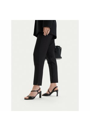 Donna che indossa pantaloni neri aderenti con spacchi alla caviglia, sandali neri con tacco alto e porta una borsa nera, in piedi su uno sfondo chiaro.