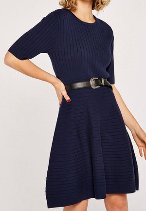 Frau trägt ein marineblaues, geripptes Strickkleid mit ellbogenlangen Ärmeln und einem schwarzen Gürtel mit silberner Schnalle, posiert mit einer Hand in der Hüfte.