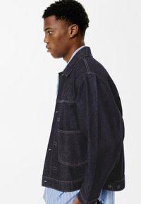 Tamna traper jakna opuštenog kroja, s velikim džepovima i vidljivim šavovima u kontrastnoj boji. Ovratnik i manžete su klasičnog dizajna.