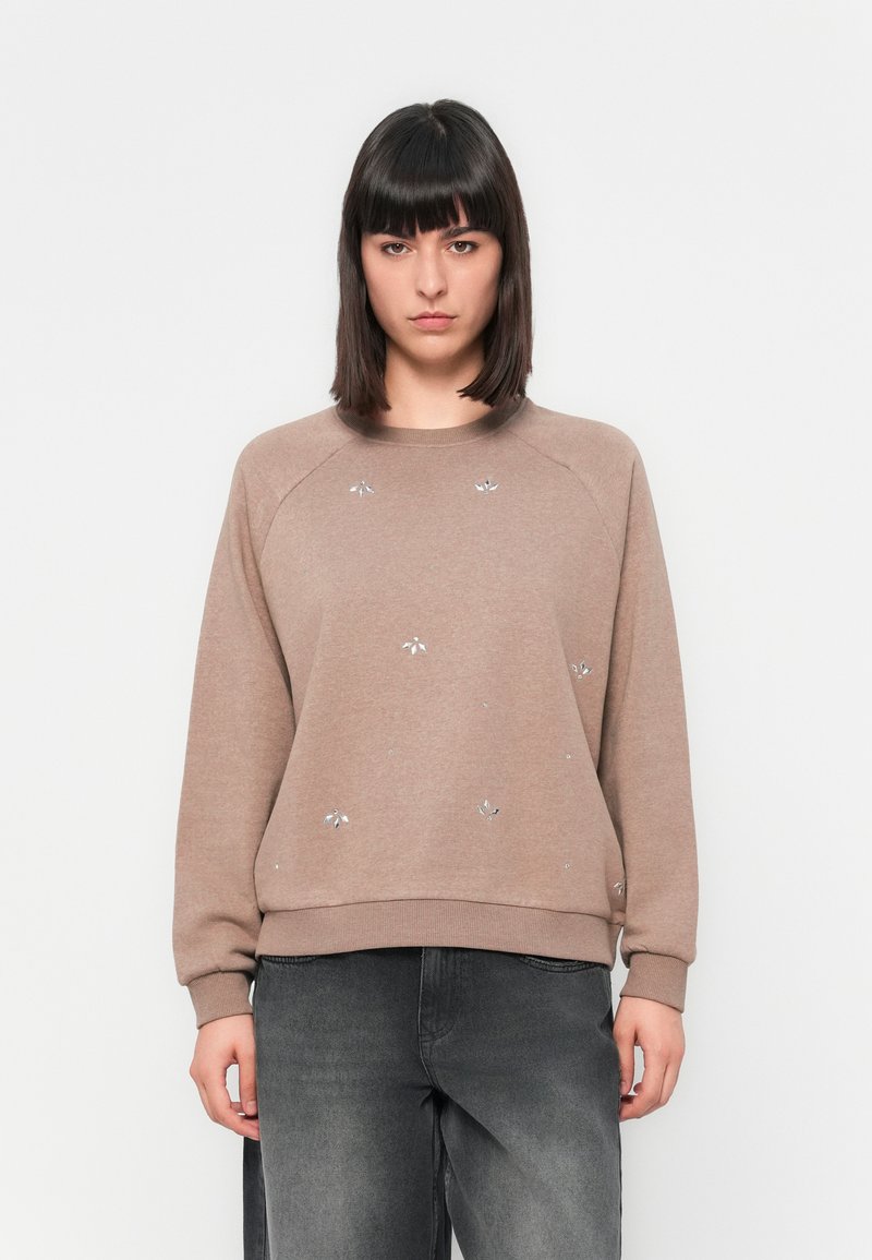 Sweatshirt marron avec un col rond et des manches raglan, orné de décorations florales argentées. Assortis avec un jean gris foncé.