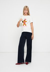 Blonde vrouw in een wit T-shirt met een feestelijke peperkoekgrafiek, marineblauwe wijde broek, rode puntige flats en een witte schoudertas.