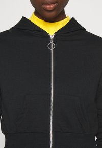 Sudadera negra con cremallera plateada, bolsillos delanteros y capucha con cordón. Llevada sobre un suéter de cuello alto amarillo. Textura de tela suave.