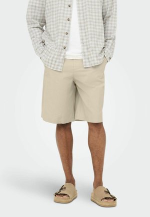 Homme portant un short beige, des sandales beige à bout ouvert avec boucles, une chemise blanche et une chemise à carreaux légère boutonnée avec les mains dans les poches.