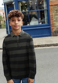 Mørkegrøn og sort stribet langærmet polo-shirt med knaplukning, iført med denim jeans, udendørs i en byindstilling.