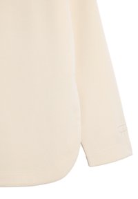 Long-sleeve shirt in off-white fabric, featuring a soft texture, rounded hem, and subtle embroidered logo near the cuff.