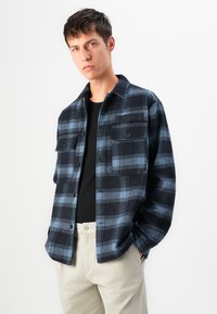 Giovane uomo che indossa una camicia a quadri blu e nera sopra una maglietta nera e pantaloni beige, in piedi contro uno sfondo bianco semplice.