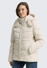 Chaqueta acolchada de color beige claro con un cuello alto, cierre frontal de cremallera y dos bolsillos. Presenta una capucha desmontable con detalles de botones.