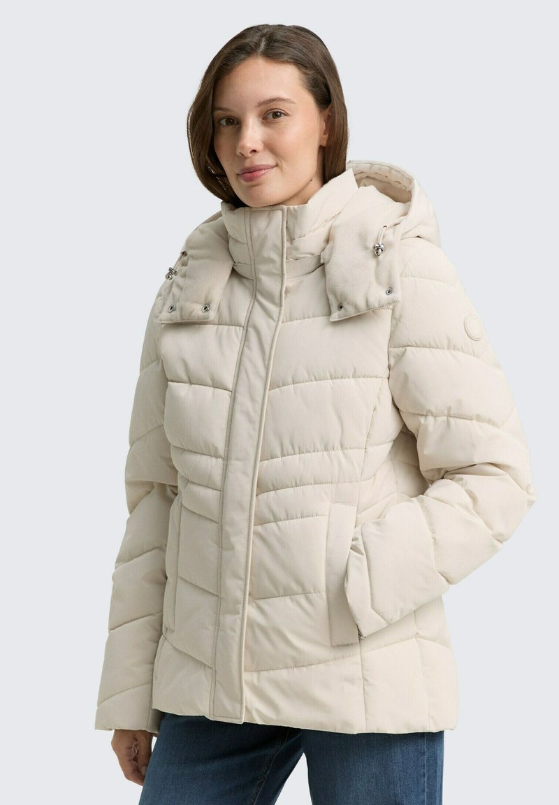 Chaqueta acolchada de color beige claro con un cuello alto, cierre frontal de cremallera y dos bolsillos. Presenta una capucha desmontable con detalles de botones.