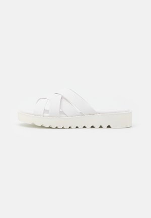 Hvid slip-in sandal med tykke krydsede remme og en chunky tekstureret sål, vist fra siden på en ensfarvet baggrund.