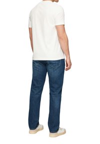 T-shirt blanc à manches courtes associé à un jean bleu. Coutures visibles et poches arrière sur le jean, design simple, et baskets beiges.