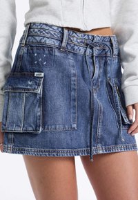 Jeansrok van blauwe stof, met voorste strik en twee grote zijzakken. Verweerd uiterlijk met lichte vervaging en subtiele markeringen.