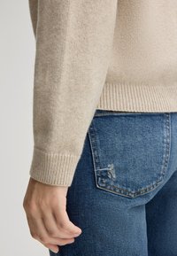 Maglione beige lavorato a maglia con polsini a costine, abbinato a jeans in denim blu con dettagli strappati sulla tasca posteriore.