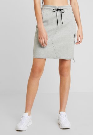 Femme portant une jupe grise à cordon avec un logo Nike noir, associée à des baskets blanches, debout devant un fond uni.