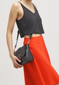 Sac à bandoulière en cuir noir avec texture crocodile, quincaillerie dorée et bandoulière réglable, tenu contre une jupe rouge vibrante.