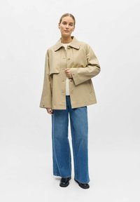 Femme debout portant une veste beige boutonnée, un pull blanc, un jean bleu à jambes larges et des chaussures noires sur un fond blanc uni.