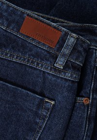 Jeans de mezclilla azul oscuro con una etiqueta de marca de cuero marrón, costuras en contraste y un bolsillo frontal con un detalle de botón de cobre.