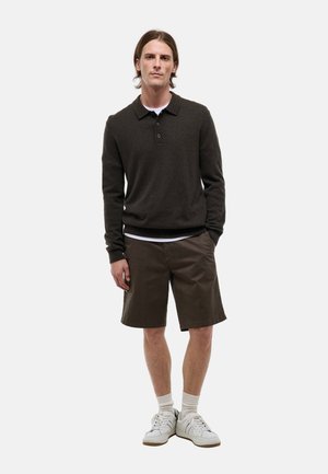 Jeune homme debout portant un pull polo à manches longues foncé, un short marron longueur genoux, des chaussettes blanches et des baskets blanches sur fond uni.