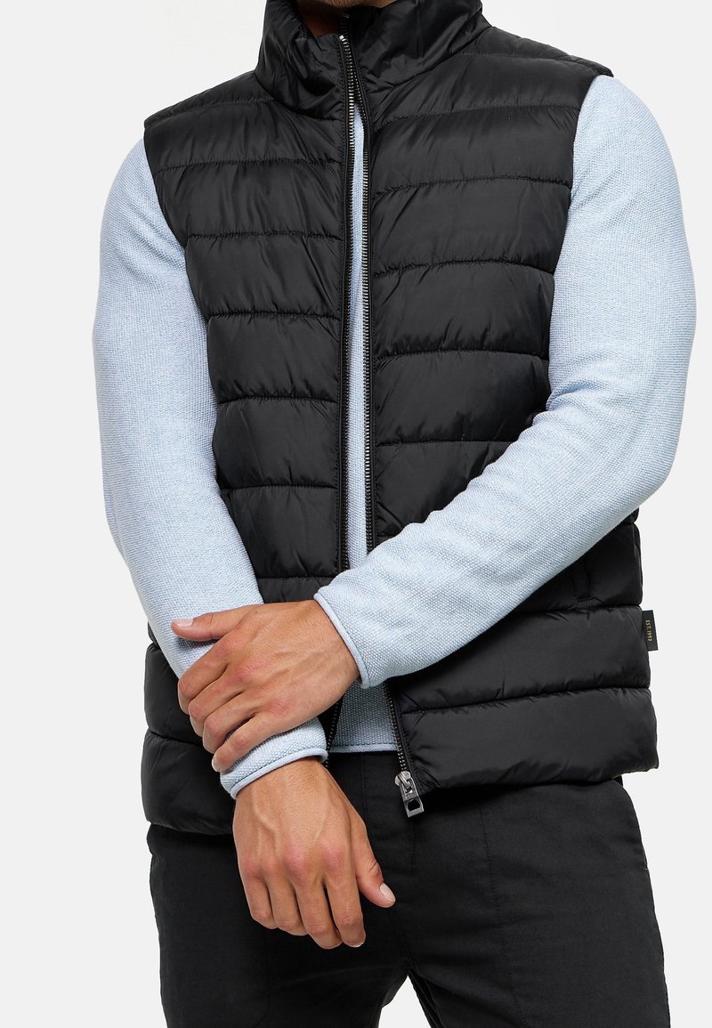 Personne portant une veste noire matelassée sans manches par-dessus un pull bleu clair à manches longues, bras croisés, debout devant un fond uni.