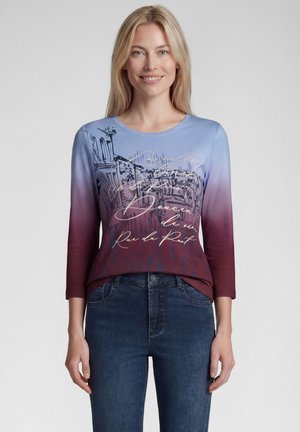 Blonde Frau trägt ein langärmliges Top mit Farbverlauf von Blau zu Burgund, mit Stadtsilhouette und kursiver Schrift, kombiniert mit dunkelblauen Jeans.