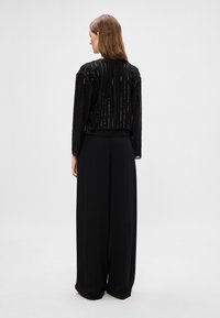 Μαύρο μπλουζάκι με μακριά μανίκια και κάθετες ραβδώσεις από φανταχτερά στρας, συνδυασμένο με μαύρες παντελόνες φαρδιάς γραμμής, από απαλό ύφασμα, χωρίς ορατά διακοσμητικά στοιχεία.