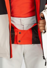 Chaqueta con un diseño en negro, blanco y rojo. Se puede ver un forro interno suave de color naranja, con un cierre a presión y una cintura elástica.