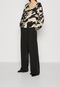 Multifarvet bluse med v-udskæring og rummelige ærmer, parret med sorte bukser med brede ben og spidse sorte sko.