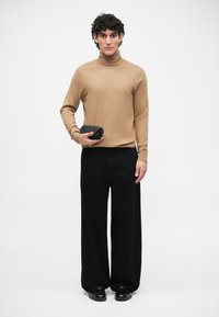 Beżowy sweter z golfem, szerokie czarne spodnie, trzymająca małą czarną torebkę. Gładka faktura, minimalistyczny design, kontrast między kolorami.