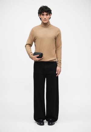 Pulover bej cu guler înalt, pantaloni negri largi, ținând o poșetă mică neagră. Textură netedă, design minimalist, contrast între culori.