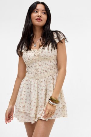 Femme aux longs cheveux foncés portant une courte robe florale crème avec des manches bouffantes, un collier pendentif, ainsi que plusieurs bracelets et bagues.