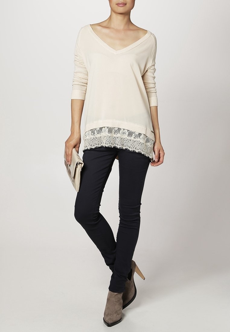 Femme portant un pull crème à col en V avec un ourlet en dentelle, un pantalon skinny noir, des bottines taupe et tenant une pochette beige.