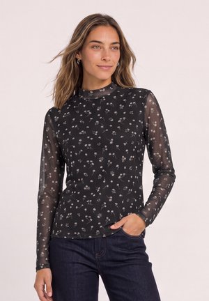 Femme aux cheveux châtain clair portant un haut noir transparent à manches longues avec un petit imprimé floral blanc et un jean bleu foncé, main dans la poche.