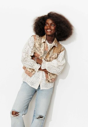 Femme souriante aux cheveux bouclés naturels portant un gilet beige à motifs, une blouse blanche à œillets, un jean bleu clair déchiré et des bijoux argentés, bras croisés.