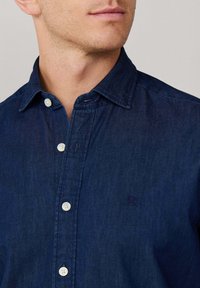 Ciemnoniebieska koszula dżinsowa z kołnierzykiem zapinanym na guziki, białymi guzikami i subtelną teksturą. Ma małe haftowane logo na piersi.