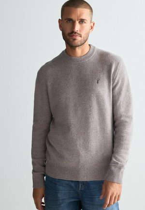 Gray knit sweater with round neckline, ribbed cuffs and hem, featuring a small embroidered accent on the chest, worn over blue jeans.