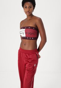 Burgundijas bezpaliku cropped tops ar logo dizainu un baltajiem akcentiem, kombinēts ar atbilstošām sarkanām sporta biksēm ar sānu svītrām un logo detaļām.