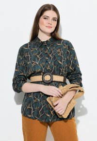 Donna che indossa una blusa scura a motivi con una larga cintura intrecciata, tiene una borsa intrecciata, abbinata a pantaloni senape, in piedi di fronte a uno sfondo bianco.