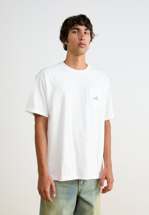 Weißes Baumwoll-T-Shirt mit Rundhalsausschnitt, kurzen Ärmeln und einer linken Brusttasche mit kleinem Logo. Kombiniert mit hellblauen Jeans.