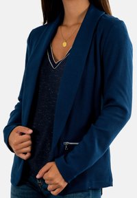Blazer azul marino con tejido texturizado, abertura frontal, cuello con muescas y detalle de bolsillo con cremallera. Usado sobre una camisa azul marino con detalles en plata.