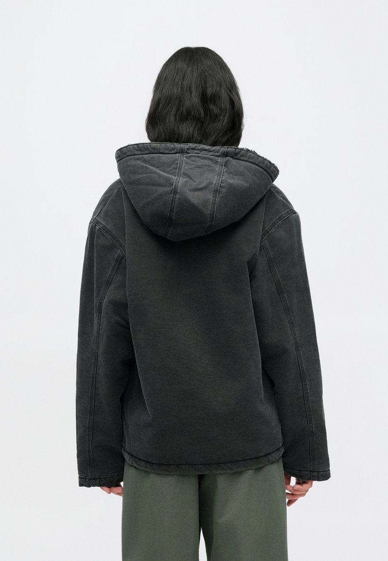 Zwarte capuchontrui van zachte stof met een oversized pasvorm, zichtbare stikdetails en een afgeronde zoom, gedragen over groene broek.