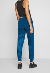 Blaue Sporthose mit seitlichen Einsätzen aus schimmerndem Material, elastischem Bund und Bündchen, kombiniert mit einem ärmellosen schwarzen Oberteil und schwarzen Schuhen.