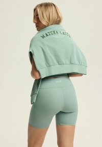 Miętowy zestaw do ćwiczeń, składający się z krótkiego swetra z napisem "MATCHA LATTE" na plecach oraz szortów o wysokim stanie z gładką teksturą.