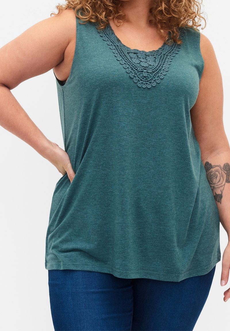 Femme portant un haut turquoise sans manches avec un détail en dentelle et un jean bleu, main sur la hanche, montrant un tatouage floral sur l'avant-bras droit.
