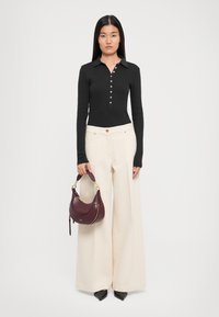 Une femme se tient face à l'avant, portant un haut noir à manches longues et boutonné, un pantalon large crème, des chaussures noires à bout pointu et tenant un sac à main bordeaux.