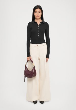 Woman stands facing forward wearing a black long-sleeve buttoned top, wide-leg cream pants, black pointed shoes, holding a burgundy handbag.