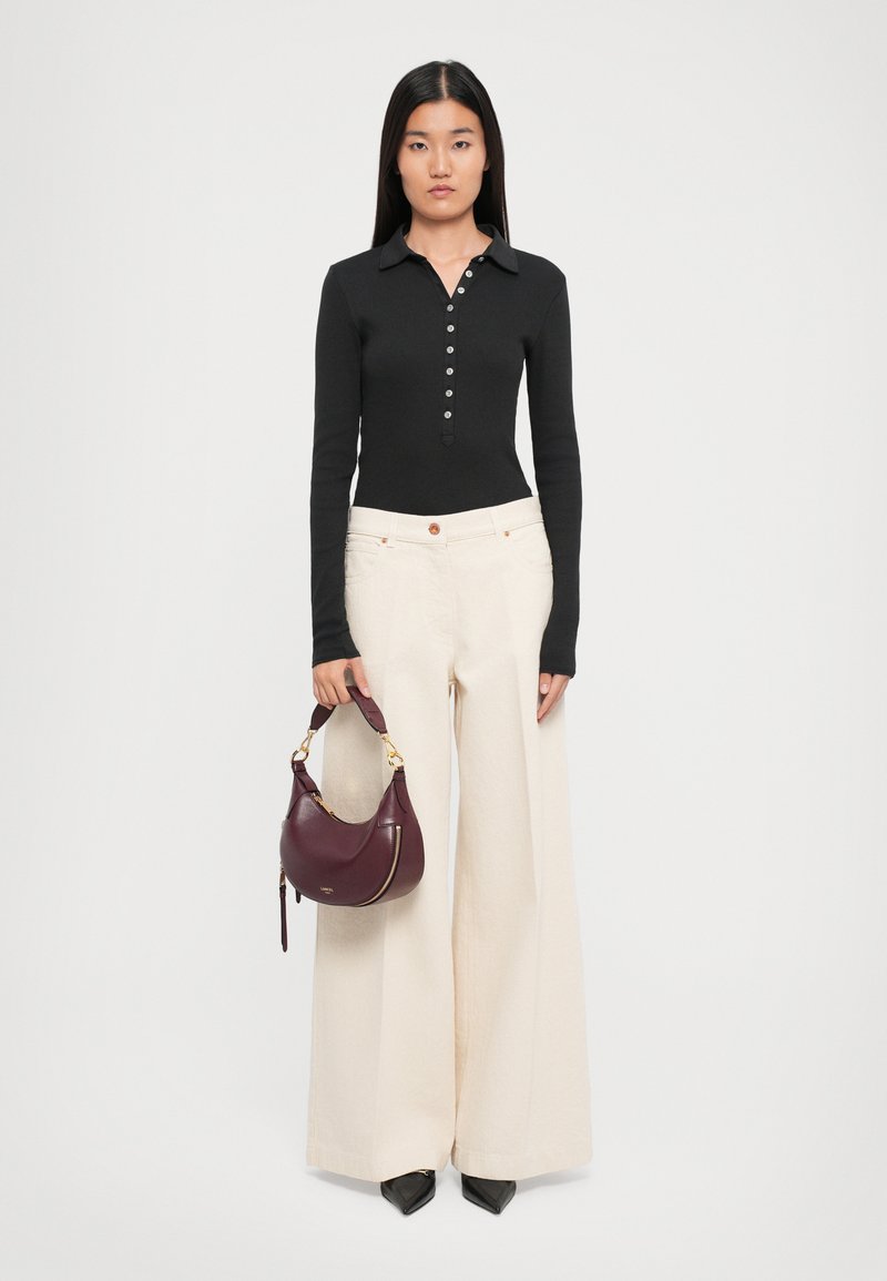 Une femme se tient face à l'avant, portant un haut noir à manches longues et boutonné, un pantalon large crème, des chaussures noires à bout pointu et tenant un sac à main bordeaux.