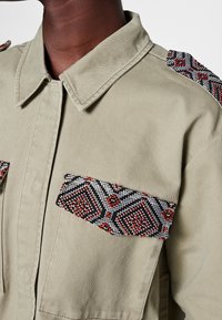 Khaki jacket featuring beaded detail on the shoulder and pocket with geometric patterns in black, red, and white. Textured fabric with a collar.