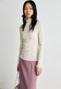 Haut à manches longues, blanc crème, avec un col à volants et une coupe ajustée, associé à une jupe rayée en diagonale dans les tons violet et rouge.