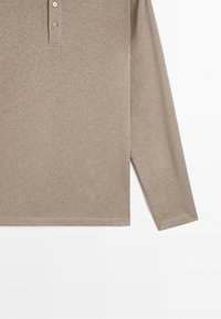 Polo beige à manches longues avec trois boutons et un ourlet droit, présenté à plat sur fond blanc.